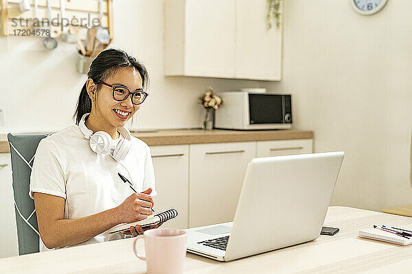 Junge Frau mit Kopfhörern  die in einen Notizblock schreibt  während sie zu Hause am Laptop arbeitet