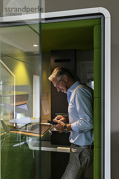Älterer Geschäftsmann  der ein Mobiltelefon benutzt  während er in einer schalldichten Kabine im Büro steht