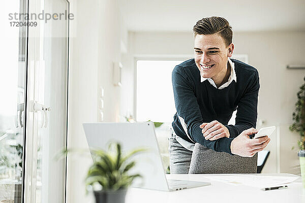 Glücklicher junger Geschäftsmann  der sich auf einen Stuhl lehnt und auf einen Laptop im Büro zu Hause schaut
