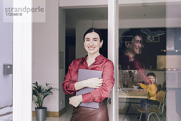 Lächelnde Geschäftsfrau  die einen Laptop hält und vor einer Glastür steht