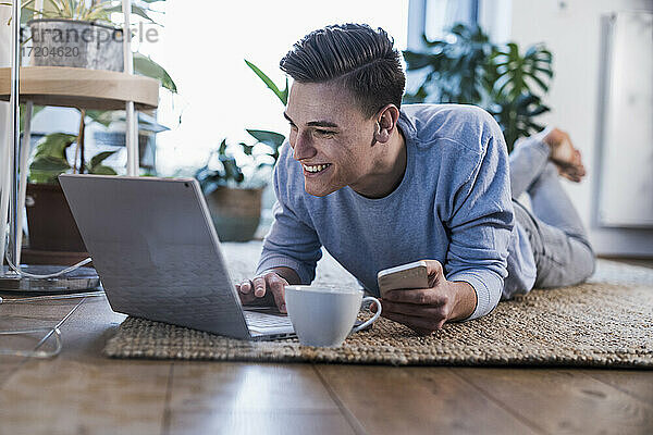 Lächelnder junger Mann mit Smartphone und Laptop  während er auf dem Teppich im Wohnzimmer liegt