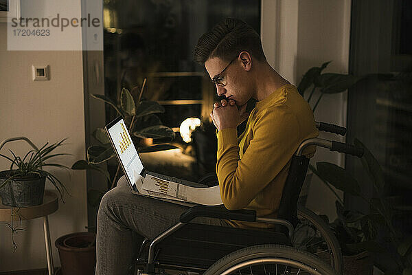 Konzentrierter junger Mann im Rollstuhl mit Laptop  der spät arbeitet  während er im Wohnzimmer im Rollstuhl sitzt