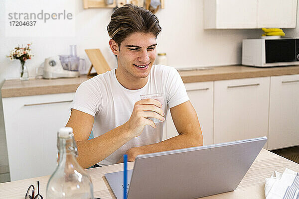 Junger Mann hält ein Glas Wasser in der Hand  während er zu Hause in der Küche am Laptop sitzt