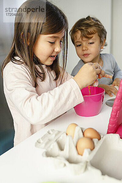 Niedlicher Junge sieht seine Schwester an  die in der Küche ein Ei in eine Schüssel schlägt