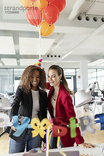 Lächelnde Geschäftsfrau hält ein Bündel Luftballons  während sie mit einer Kollegin im Büro Geburtstag feiert