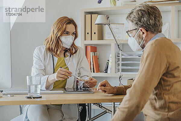 Arzt mit Gesichtsschutzmaske bei der Erläuterung von Medikamenten in einer Arztpraxis