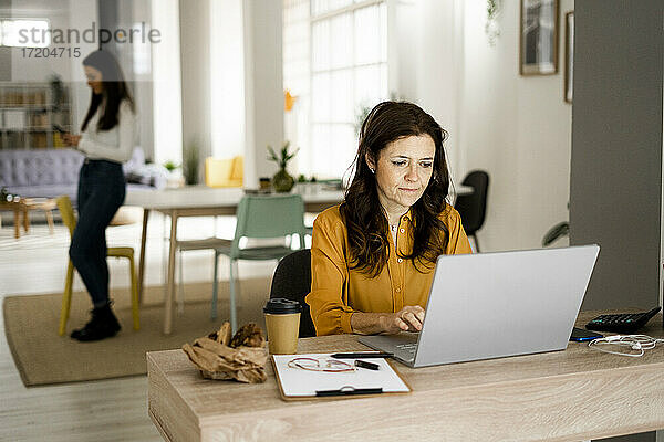 Reife Geschäftsfrau  die an einem Laptop arbeitet  während ihre Tochter im Büro zu Hause ein Mobiltelefon benutzt