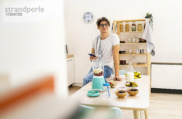 Junger Mann mit Smartphone sitzt auf dem Tisch in der Küche zu Hause