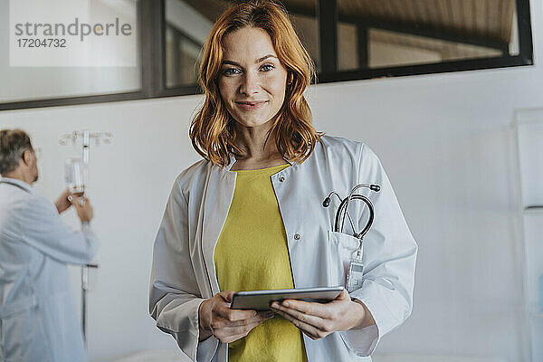 Eine Ärztin hält ein digitales Tablet  während sie mit einem Mitarbeiter im Hintergrund in einer Klinik steht