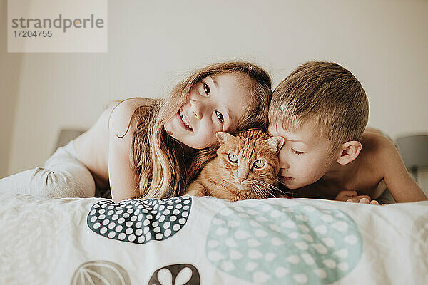 Lächelndes Mädchen mit Bruder  der sich auf eine Katze im Schlafzimmer stützt  zu Hause