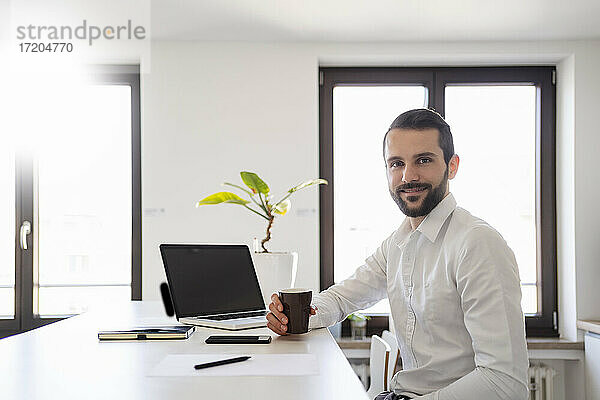 Lächelnder junger Geschäftsmann mit Kaffeetasse  der im Büro am Laptop sitzt