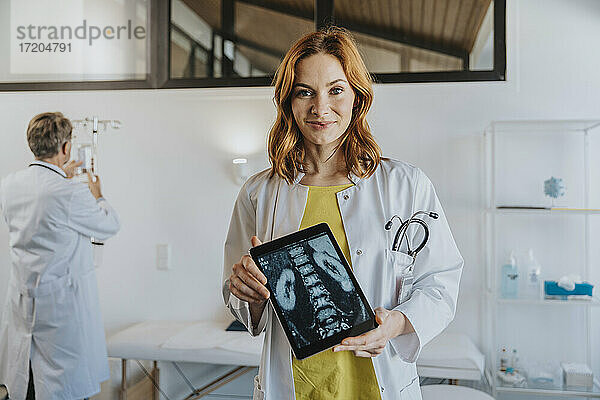 Arzt zeigt Röntgenbild auf digitalem Tablet  während er mit einem Mitarbeiter im Hintergrund in einer Klinik steht