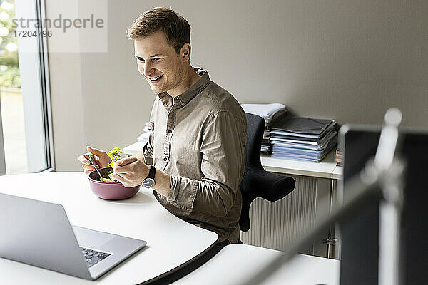 Lächelnder junger Geschäftsmann beim Mittagessen mit Blick auf den Laptop im Büro
