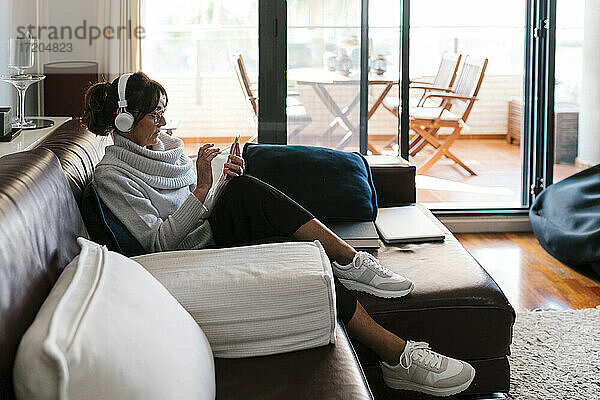 Reife Frau mit Kopfhörern  die ein Mobiltelefon benutzt  während sie zu Hause sitzt