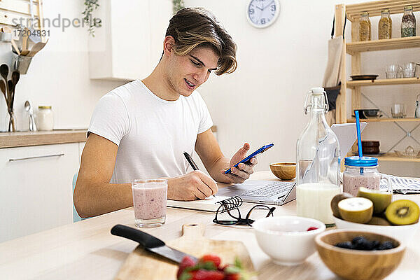 Junger Mann mit Smartphone  der auf einem Notizblock schreibt  während er am Tisch in der Küche sitzt