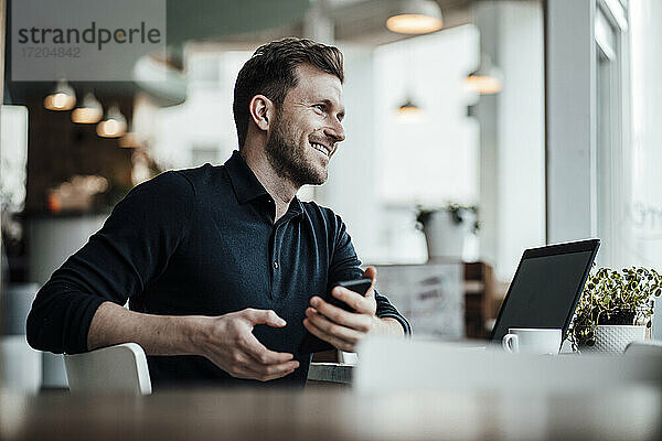 Lächelnder Geschäftsmann mit Laptop und Mobiltelefon  der in einem Café sitzt und wegschaut