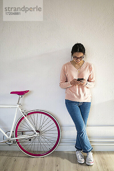 Lächelnde Frau  die ein Mobiltelefon benutzt  während sie zu Hause am Fahrrad steht
