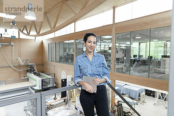 Lächelnde Geschäftsfrau mit digitalem Tablet  die sich an ein Geländer in einer Fabrik lehnt