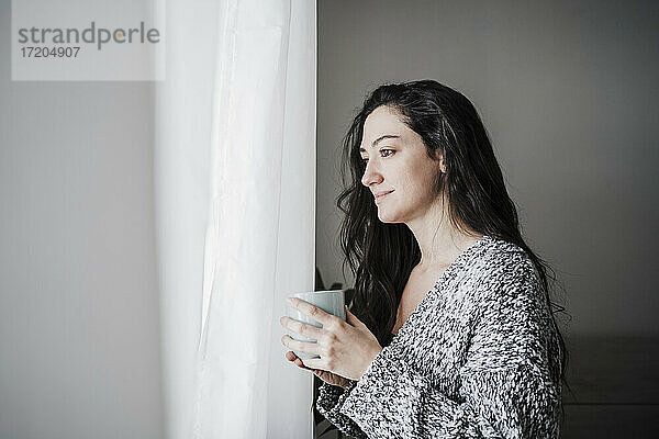 Nachdenklich lächelnde Frau  die eine Kaffeetasse hält  während sie durch ein Fenster zu Hause schaut