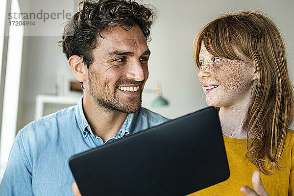 Lächelnde Tochter sieht ihren Vater an  während sie zu Hause ein digitales Tablet hält