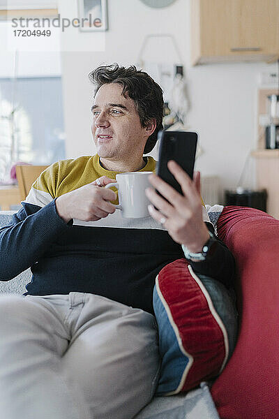 Mann mit Smartphone und Kaffeetasse auf dem Sofa sitzend und wegschauend