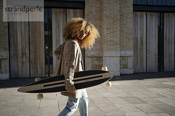 Afro-Frau  die ein Skateboard trägt  während sie an einem sonnigen Tag auf dem Fußweg gegen ein Gebäude läuft