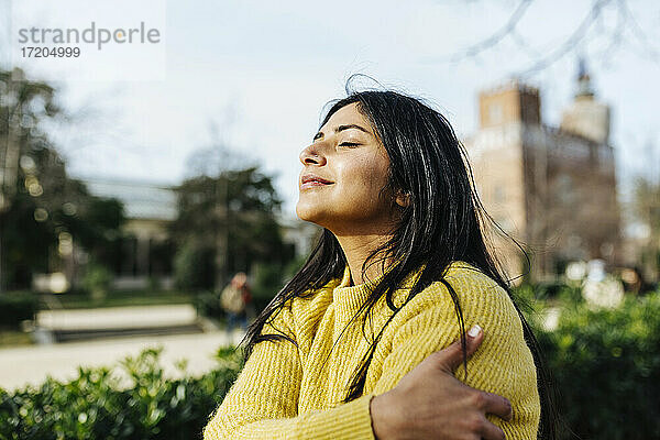 Lächelnde Frau umarmt sich selbst gegen den Himmel in einem öffentlichen Park