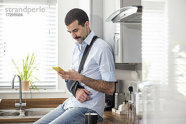 Ein verletzter Mann trägt eine Armschlinge  während er auf dem Küchentisch zu Hause sitzt und ein Mobiltelefon benutzt