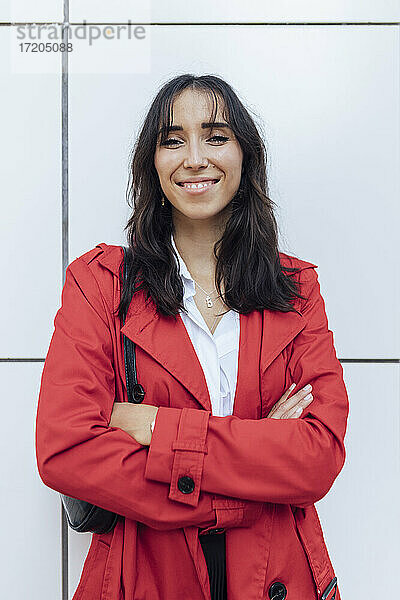 Lächelnde junge Frau in rotem Trenchcoat steht mit verschränkten Armen vor einer weißen Wand