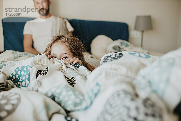 Mädchen versteckt sich unter einer Decke  während der Vater zu Hause auf dem Bett zusieht