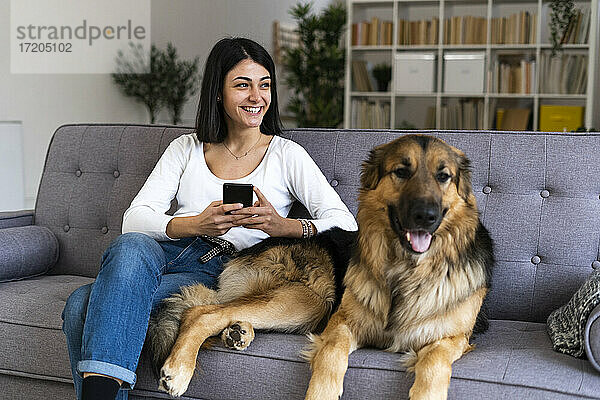 Glückliche Frau mit Smartphone  die wegschaut  während sie mit ihrem Hund zu Hause auf dem Sofa sitzt