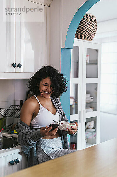 Lächelnde Frau  die eine Obstschale hält  während sie ihr Smartphone in der Küche benutzt