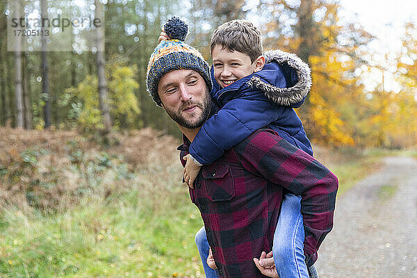 Lächelnder Junge genießt es  im Wald huckepack auf seinem Vater zu sitzen