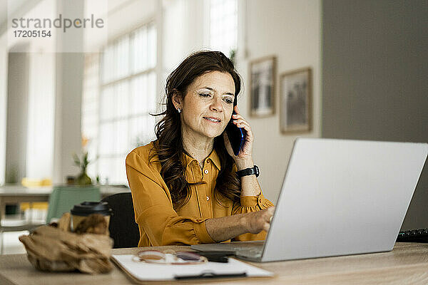 Lächelnde Geschäftsfrau  die einen Laptop benutzt  während sie am Schreibtisch im Heimbüro mit dem Handy telefoniert