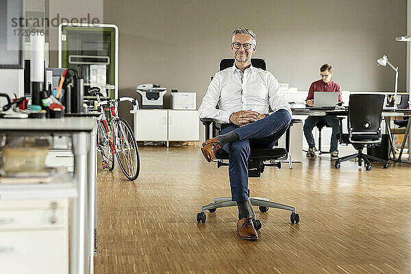 Lächelnder Geschäftsmann mit gekreuzten Beinen auf einem Stuhl im Büro sitzend