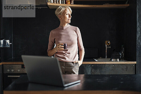 Unternehmerin mit den Händen in den Taschen  die wegschaut  während sie in der Küche im Büro zu Hause Kaffee trinkt