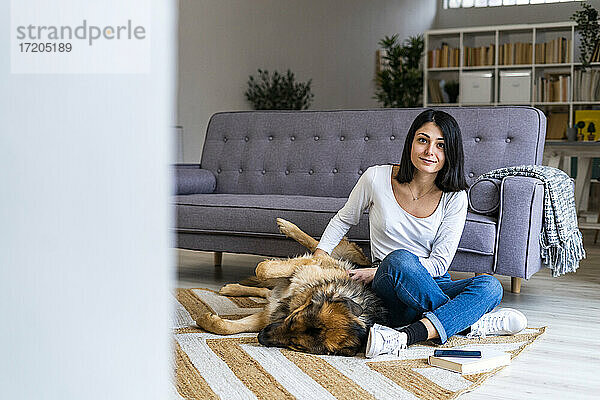 Junge Frau  die ihren Hund streichelt  während sie auf dem Teppich vor dem Sofa im Wohnzimmer sitzt