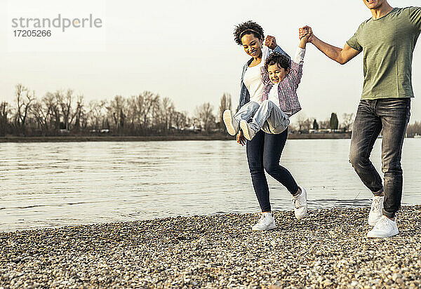 Fröhlicher Junge schaukelt mit Unterstützung seiner Eltern über einen Kieselstein am See