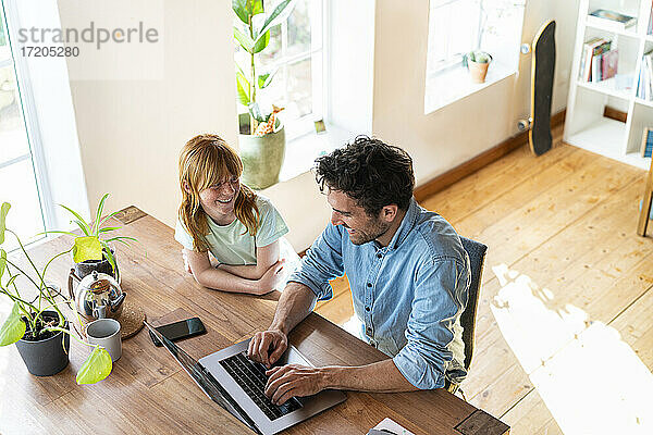 Lächelndes Mädchen  das seinen Vater bei der Arbeit am Laptop im Wohnzimmer beobachtet