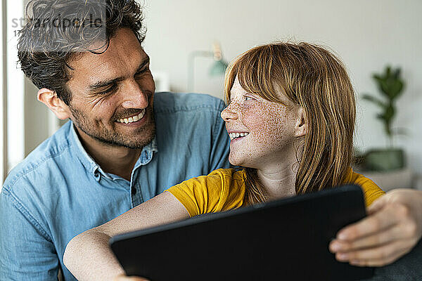 Vater und rothaarige Tochter mit digitalem Tablet lächelnd  während sie sich gegenseitig im Wohnzimmer anschauen
