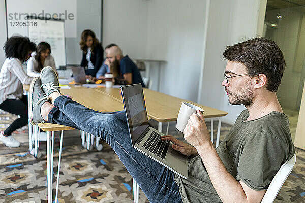 Männlicher Unternehmer hält eine Tasse  während er während einer Besprechung im Büro am Laptop arbeitet