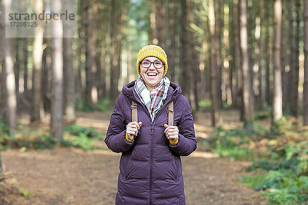 Glückliche Frau in warmer Kleidung  die einen Rucksack hält  während sie im Wald steht