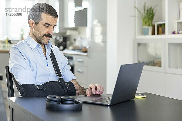 Mann mit Armschlinge arbeitet zu Hause am Laptop