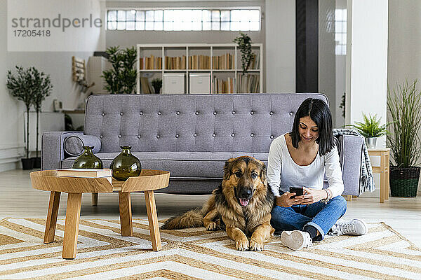 Frau  die ein Mobiltelefon benutzt  während sie mit ihrem Hund im Wohnzimmer auf dem Teppich sitzt