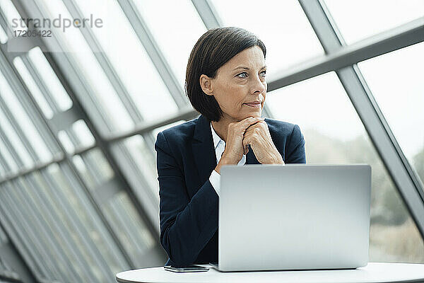 Nachdenkliche Geschäftsfrau mit Hand am Kinn neben dem Laptop am Schreibtisch im Büro