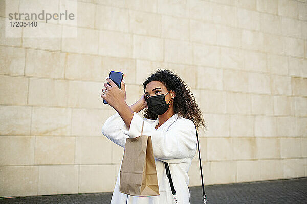 Junge Frau  die ein Selfie mit ihrem Smartphone auf dem Fußweg vor einer Mauer macht