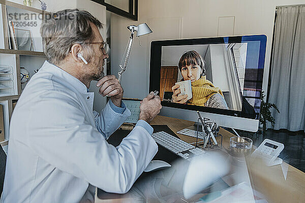 Älterer Arzt im Gespräch mit einem Patienten per Videoanruf über einen Computer  während er in der Arztpraxis sitzt