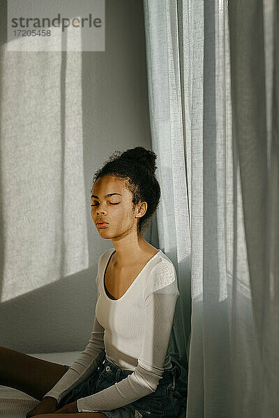 Junge Frau mit geschlossenen Augen sitzt zu Hause vor einem Vorhang