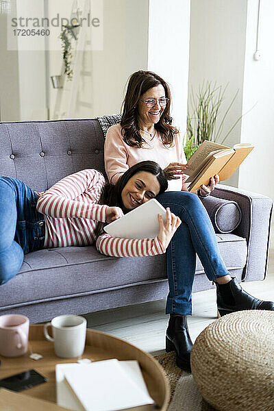 Lächelnde Tochter  die ein digitales Tablet benutzt  während sie auf dem Schoß der Mutter liegt und ein Buch auf dem Sofa zu Hause liest