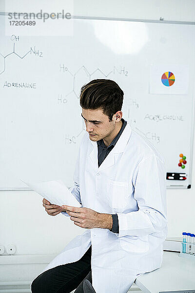 Junger männlicher Wissenschaftler schaut auf eine Tabelle  während er auf einem Schreibtisch im Labor sitzt